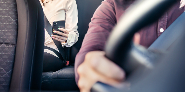 Un passager, attaché par sa ceinture de sécurité, est assis sur la banquette arrière d'une voiture de covoiturage, un téléphone intelligent à la main, tandis que le conducteur tient le volant depuis le siège avant.