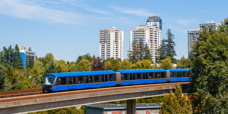 Une rame du SkyTrain de Vancouver circule sur une voie surélevée au-dessus d'un quartier arboré, parsemé d'immeubles résidentiels de grande hauteur, sous un ciel bleu limpide.