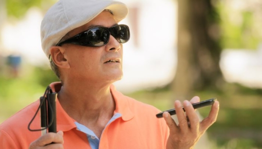 A man wearing sunglasses and a hat holds an iPhone in his left hand. A white cane sits in his right hand.