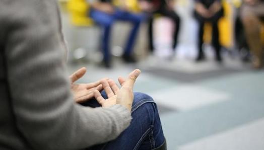 A person sits in a group circle. Their hands are clasped in their lap.