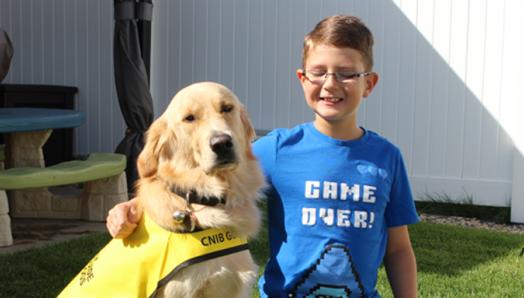 Un garçon souriant portant des lunettes avec son bras autour d'un Golden Retriever qui porte une veste jaune.