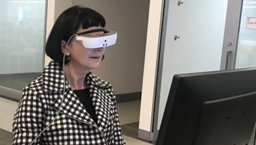 A woman in a black and white checkered jacket, wears white eSight glasses and works at her computer.