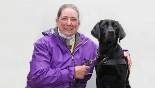 A woman in a purple jacket and her black Lab/Golden Retriever cross guide dog.