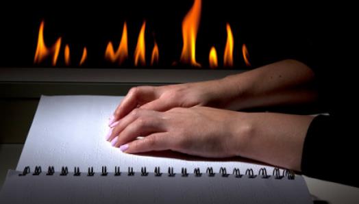 Des mains se déplacent sur un livre en braille. Une cheminée émet des flammes en arrière-plan.