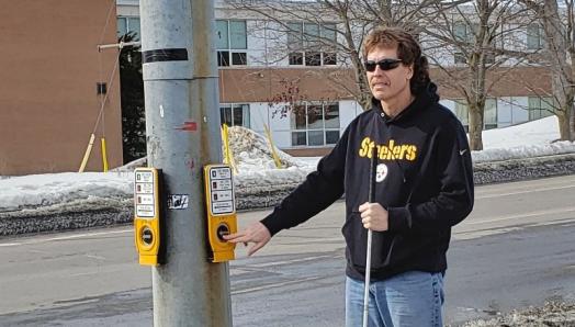 Bill Liggins se tient au coin de la rue à un panneau de signalisation piétonnière accessible. Il tient sa canne blanche dans la main gauche et appuie sur le bouton du passage pour piétons avec la main droite.