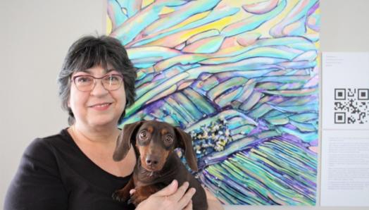 Robyn Rennie pose pour une photo devant son œuvre d'art colorée à côté d'un code QR imprimé numériquement. Elle tient dans ses bras un petit chien brun chocolat.