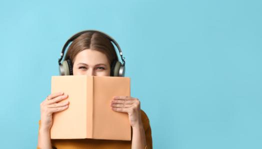 A young woman wears a pair of headphones and holds a book up to her face. The book is blocking her nose and mouth so only her eyes appear.