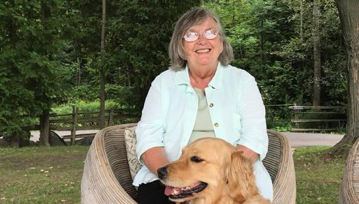 Photo de Claire assise sur une chaise à l'extérieur et souriant pour la caméra. Un golden retriever est assis à ses pieds.