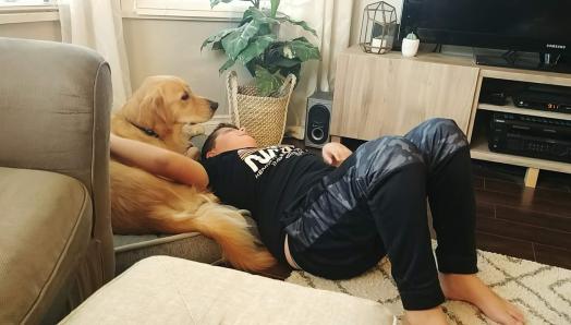 Landon and his CNIB Buddy Dog Ruggles, a Golden Retriever, laying on a cushion together in their living room. Landon’s arm is around Ruggles, petting his back.