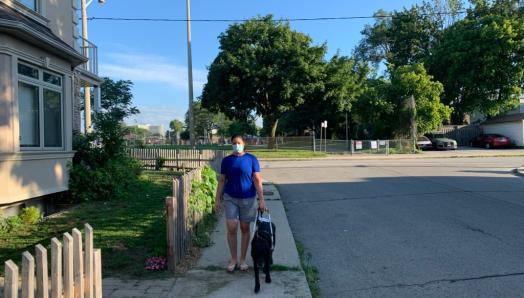 Victoria Nolan and her guide dog, Alan, walking along a sidewalk on a sunny day. Victoria is wearing a face mask.