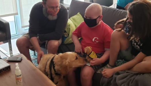 Ollie and family members sitting on a couch wearing face masks. Ambassador Dog Ziggy is sitting on the floor in front of Ollie, sniffing the stuffed CNIB Guide Dog plush puppy on Ollie's lap.