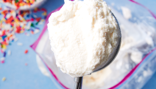 Close of up a silver ice cream scoop with a scoop of vanilla ice cream above a zip lock bag of ice cream with colourful sprinkles in a bowl on the left.