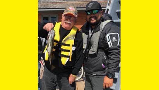 Bruce Roulston posant à côté de Eugene Chong avec un poisson pêché du quai sur la rive du Lake Joseph. Ils portent tous deux des gilets de sauvetage et une casquette.
