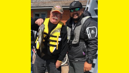 Bruce Roulston posing with a caught fish on a dock beside Eugene Chong at CNIB Lake Joe. They are both wearing life jackets and a ballcap.