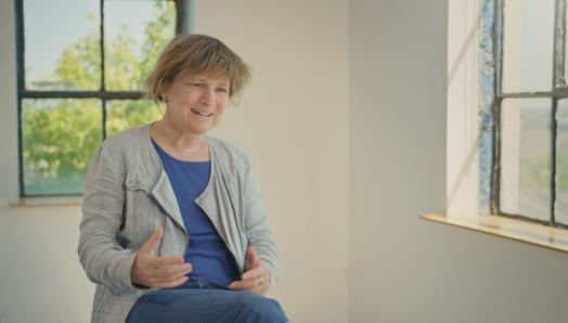 Anne Jarry sitting on a stool in a sunlit room, speaking for the camera.