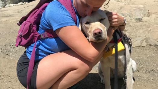 Sara, wearing hiking clothes, kneeling down giving Daisy a hug on a sunny day.