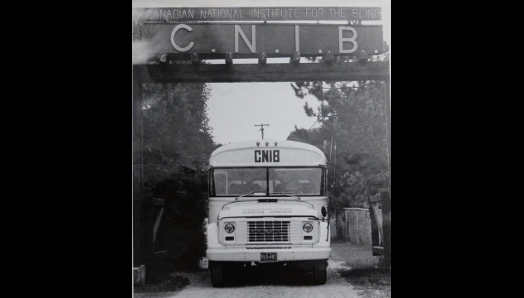 Une photo en noir et blanc d'un ancien autobus scolaire bien marqué CNIB à l'avant, passant sous le panneau de bienvenue au Centre Lake Joseph d’INCA.