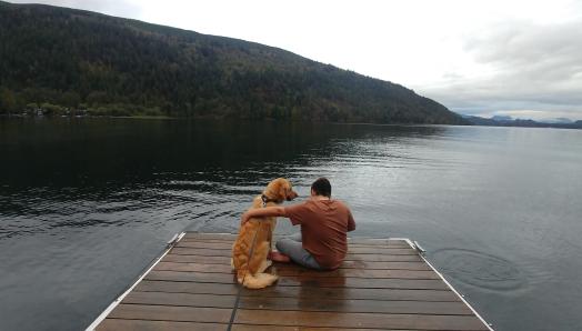 Landon and Ruggles, a golden retriever, sitting on a wharf overlooking a lake; Landon’s arm is around Ruggles who is looking at Landon.