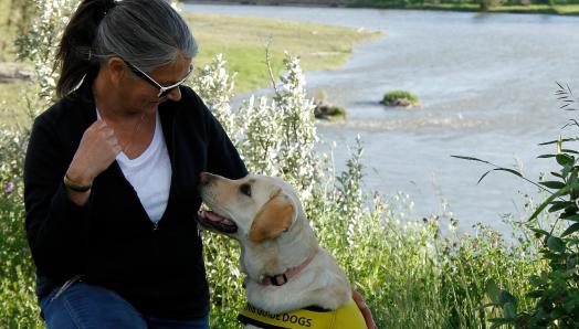 Debra Williamson agenouillée à côté de Patsy, un Labrador-Retriever blond portant un gilet de futur chien-guide, qui s’échangent un regard devant la Bow River à Calgary