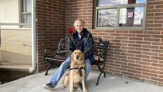 Photo de Terry assis dehors sur un banc en métal, souriant pour la caméra avec Bert assis devant lui.