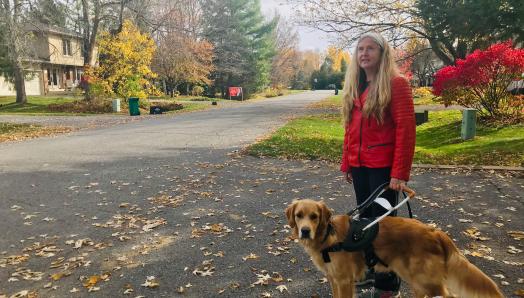 Diane, vêtue d’une veste rouge, est debout dans une rue couverte pleine de feuilles d'automne multicolores, tenant le harnais de Carla et souriant pour la pose.