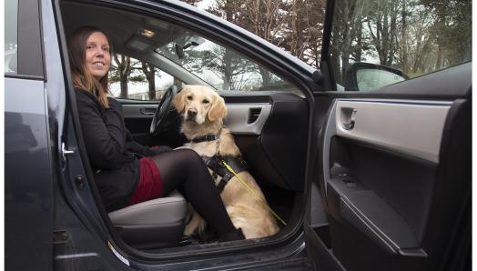 A woman sitting in the front passenger seat of a taxi with her golden retriever guide dog sitting on the floor at her feet with the car door open.