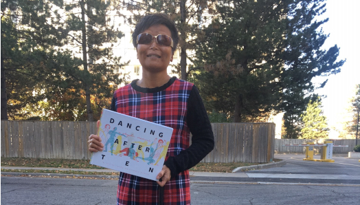 Vivian Chong se tient à l'extérieur et tient son livre, Dancing after TEN. Son chien guide noir, Catcher, se tient à ses pieds. Tout sourire, Vivian porte une robe à carreaux et des lunettes de soleil.