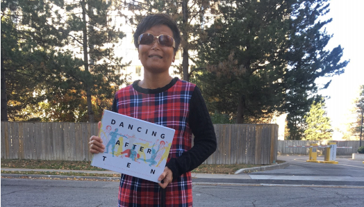 Vivian Chong se tient à l'extérieur et tient son livre, Dancing after TEN. Son chien guide noir, Catcher, se tient à ses pieds. Tout sourire, Vivian porte une robe à carreaux et des lunettes de soleil. 