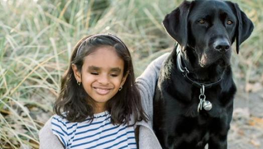 Deepa and Chelsey, a black Labrador-Retriever, sitting on sand in front of a grassy area; Deepa’s arm is around Chelsey and she is smiling for the camera.