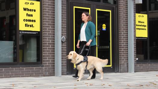 Une femme souriante qui marche avec son chien-guide, un Golden Retriever, le long d'un trottoir devant un hub communautaire d'INCA.