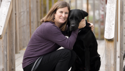 Photo of Shelley sitting on a wooden porch, smiling while giving Rookie a big hug. 