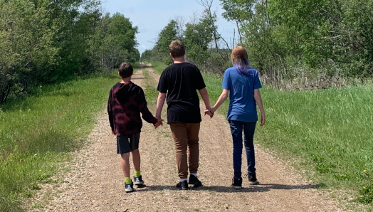 Les trois enfants d'Ashley vus de dos alors qu'ils marchent le long d'une route isolée dans une zone verte et luxuriante. Les enfants se tiennent la main. 
