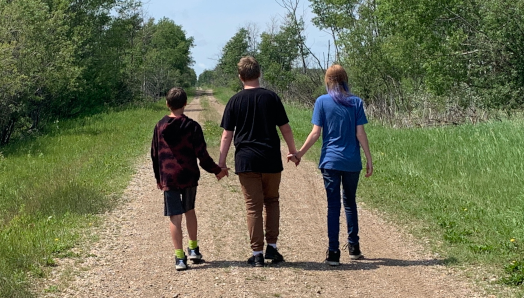 Les trois enfants d'Ashley vus de dos alors qu'ils marchent le long d'une route isolée dans une zone verte et luxuriante. Les enfants se tiennent la main.