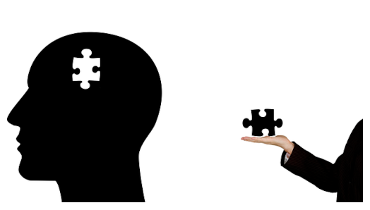 An illustration/silhouette of a bald head. A puzzle-piece is missing from inside the head. To the right of the head, a stranger’s hand extends from the frame holding the missing puzzle piece. 