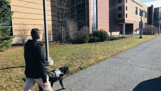 Un jeune homme marchant sur un trottoir avec son chien-guide, un croisement de Labrador-Retriever noir aux pattes bringées