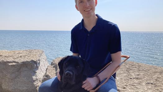 Jack McCormick and his guide dog, Baloo, a black Labrador retriever, sitting on large rocks in front of the ocean and smiling for the camera