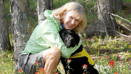 Cheryl entourée d'arbres et de verdure dans une forêt de Canmore, en Alberta, accroupie au côté d'Irwin, un labrador retriever noir portant un gilet jaune Futur chien-guide.