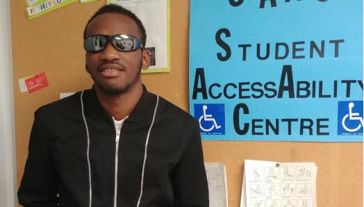  Eitel Houedakor smiling and standing in front of a bulletin board. He is wearing sunglasses and a hoodie.