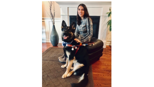 Stela sitting on a leather ottoman in front of a fireplace with her German Shepherd guide dog in front of her.