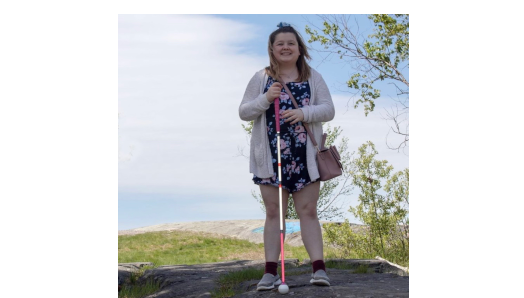 Alicia Chenier smiles and poses for a photo outdoors. Lush green trees and a blue sky in the background. She holds her white cane in her right hand.