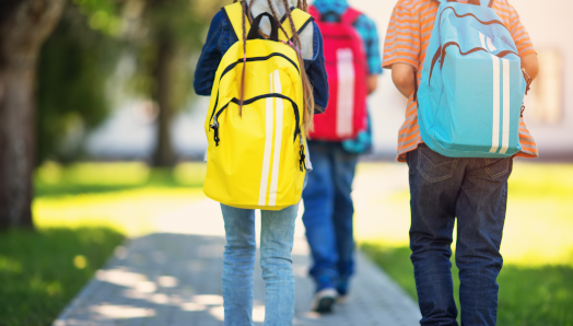 : The backs of three youth walking to school. They are wearing backpacks and are outdoors.