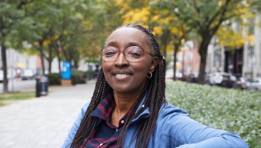 Binta is sitting on an outdoor wooden bench. She is smiling and wearing glasses.