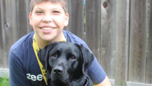 Connor is looking at the camera and has his arms around June, giving her a big hug; June, a black lab, is sitting looking at the camera.