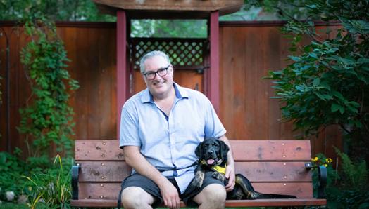 Christopher Adams sitting on a bench with a very young Percy sitting beside him wearing his bright yellow Future CNIB Guide Dogs vest.