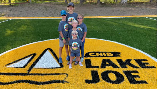 Five children posing on the giant yellow CNIB Lake Joe logo in the middle of the new soccer field. They are all wearing grey CNIB Lake Joe tshirts.