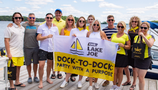 CNIB Muskoka Dock-to-Dock event volunteers posing at the CNIB Lake Joe boathouse, holding event flags.