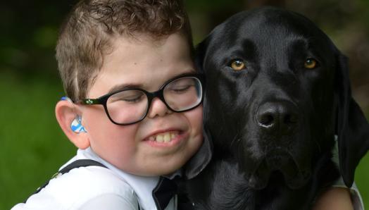 Kaiden has his eyes closed and is leaning into Flinn and giving him a big hug;  Flinn, a black lab, is sitting looking at the camera.