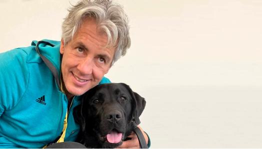 A picture of Ary wearing a turquoise Adidas jacket and black pants, sitting on the floor giving Don, a CNIB Guide Dog, a belly rub while smiling for the camera.