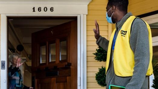 A CNIB Fundraiser in a yellow vest and face mask waving at a donor as she closes her front door