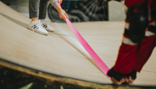 Two skateboarders place bright pink tape down on a skatepark ramp to create a more tactile/accessible skateboarding experience.